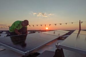 Man installs commercial solar panels on a roof with sun setting in background