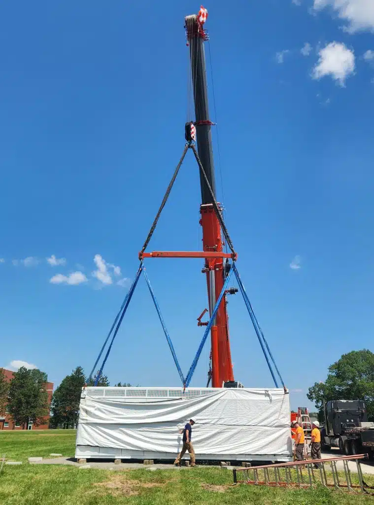 A construction crane lowers a large Tesla battery pack onto a concrete pad